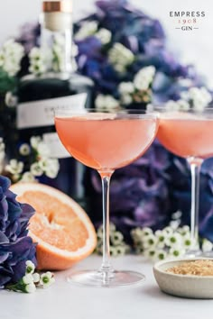 two wine glasses filled with pink liquid next to grapefruits and an orange slice