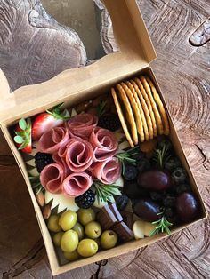 an open box filled with assorted food items on top of a wooden table next to crackers and olives