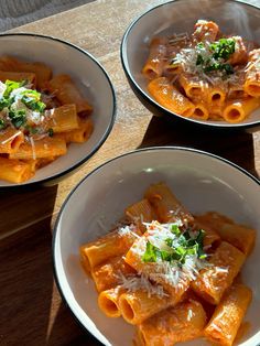 three white bowls filled with pasta and sauce on top of a wooden table in front of a window