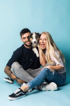 a man and woman sitting on the ground with a dog in their lap, smiling at the camera