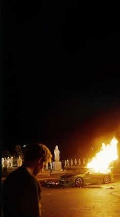 a man standing next to a car on a street with a fire in the air