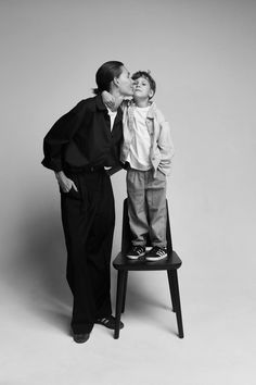 a woman kissing a young boy on the cheek while standing on a chair with his legs crossed