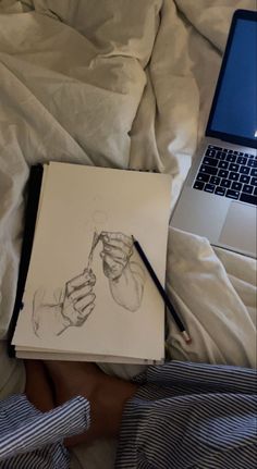 a person laying in bed next to a laptop computer and drawing something on paper with a pencil