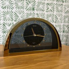 a clock sitting on top of a wooden table next to a wallpapered wall