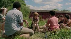 a group of people sitting on the ground in front of a red building and some tall grass