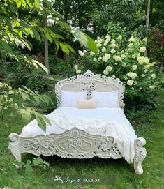 a white bed sitting in the middle of a lush green field next to trees and bushes