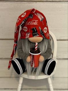 a stuffed toy sitting on top of a chair with a cup in its mouth and a coca - cola hat