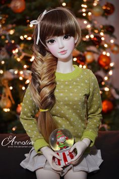 a doll with long hair sitting in front of a christmas tree wearing a green sweater