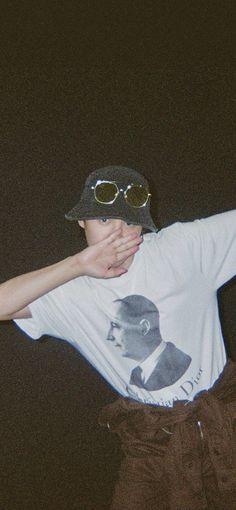 a young man wearing sunglasses and a hat covers his face with his hands while standing in front of a dark background