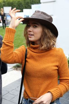 a woman in an orange sweater and brown hat is holding her hand up to the side