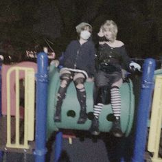 two people sitting on top of a green and yellow playground structure with blue poles, wearing face masks