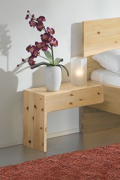 a white vase with purple flowers sitting on a small wooden table next to a bed