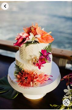 a three tiered cake with flowers on it sitting on a table next to the ocean