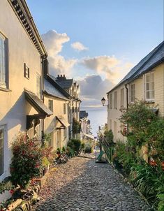 a cobblestone street with houses and flowers on both sides, leading to the ocean