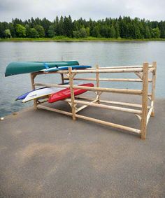 Freestanding log kayak rack on a concrete boat ramp next to a lake.  There are several kayaks and a canoe on the rack.  Several trees surround the lake in the background. Wood Kayak Rack Plans, Landscaping Blocks, Wood Kayak, Dock House, Water Vessel, Northern White Cedar, Kayak Storage, Cedar Log, Kayak Rack