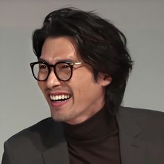 a man wearing glasses and a black turtle neck sweater smiles at the camera with his right hand on his hip