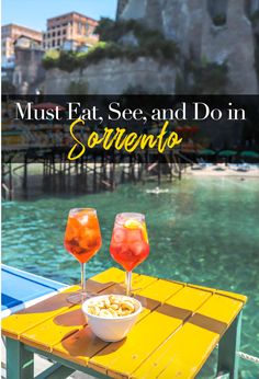 two glasses of wine sitting on top of a yellow table next to the ocean with text overlay that reads must eat, see and do in sorrento