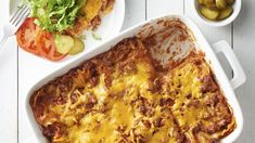 a casserole dish with meat, cheese and vegetables next to it on a white table