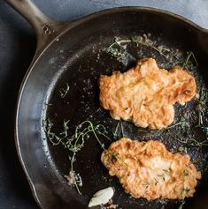 two fried meat patties in a frying pan with herbs on the side,