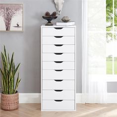 a white dresser with black drawers in front of a gray wall and potted plant