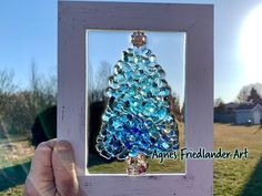 a hand holding up a framed glass christmas tree ornament in front of a grassy field