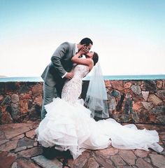 a bride and groom kissing in front of a stone wall with the ocean in the background