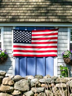 an american flag hanging on the side of a house with flowers in front of it