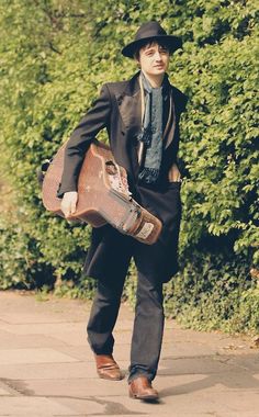 a man in a suit and hat carrying a skateboard down a sidewalk with bushes behind him