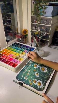 a person is painting flowers with watercolors on a table in front of a window