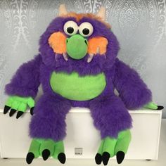 a purple and green monster stuffed animal sitting on top of a white box with black feet