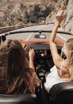 two women are riding in the back of a convertible car on a mountain road and one is holding her hand up