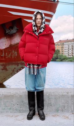 a man standing in front of a boat wearing a red puffy jacket and black boots