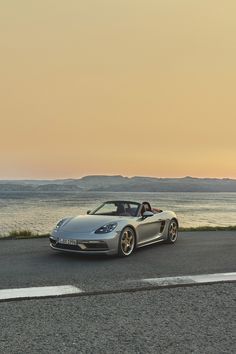 a silver sports car parked on the side of the road next to the ocean at sunset