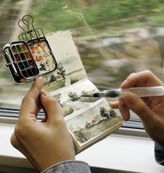 a person holding an open book next to a window with watercolors on it