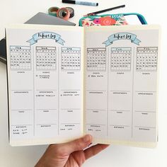 a hand holding an open planner in front of a white table with pens and pencils