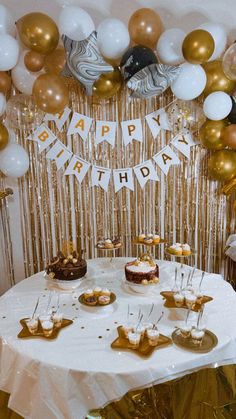 a white table topped with lots of cake and desserts covered in gold foil balloons