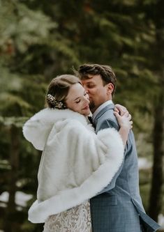 a bride and groom embracing in the snow surrounded by pine trees at their winter wedding