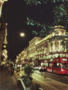 a red double decker bus driving down a street next to tall buildings at night time