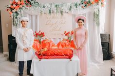 two people standing in front of a table with flowers and decorations on the wall behind them