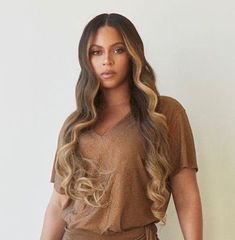 a woman standing in front of a white wall wearing a brown shirt and tan pants