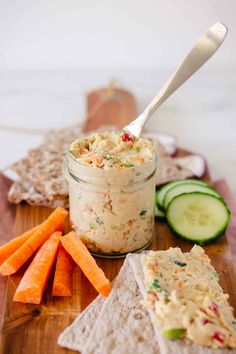 a wooden cutting board topped with crackers and veggies next to a jar of dip