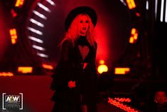 a woman in a black outfit and hat on stage with red lights behind her at a concert