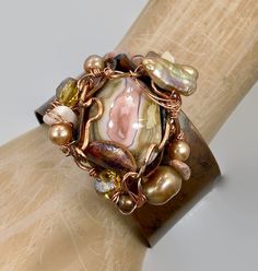 "Pink and shades of soft browns and beige....  Starting with a copper cuff bracelet that I have hammered and heated for an amazing color... I then chose the large jasper stone that is the centerpiece of this bracelet. I love the swirling shades of pink and cream.  I encased it in a freeform, forged copper bezel with a unique organic shape to it.  I have given it an artistic and organic wire wrap . For additional accents I have used beads in stone, glass and pearls to enhance the shades of the st Bohemian Adjustable Copper Bracelets, Elegant Brown Cuff Bracelet As Gift, Elegant Brown Cuff Bracelet Gift, Hand Wrapped Brown Copper Bracelets, Hand Wrapped Copper Bracelets In Brown, Brown Hand Wrapped Cuff Bracelet For Gift, Adjustable Brown Copper Bangle, Adjustable Hand Wrapped Bronze Cuff Bracelet, Bohemian Copper Bangle For Gifts