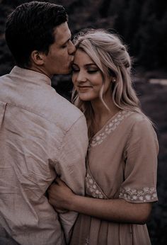 a man and woman standing next to each other in front of some trees with their faces close together