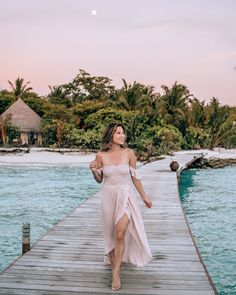 a woman in a dress walking on a dock near the ocean with palm trees behind her