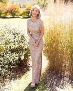 a woman wearing a gold dress standing in front of some tall grass and bushes with her hand on her hip