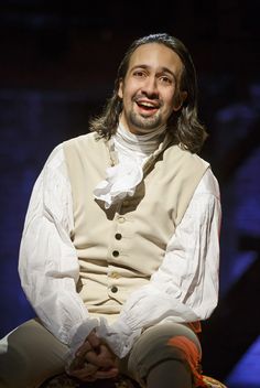 a man with long hair wearing a white shirt and vest sitting on a chair smiling at the camera