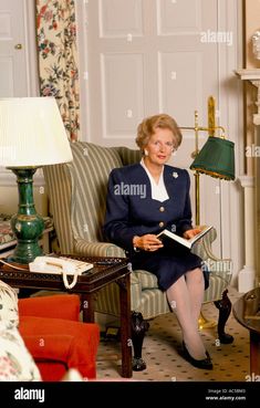 an older woman sitting on a chair in her living room reading a book - stock image