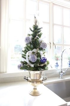 a small christmas tree in a silver pot next to a kitchen sink and window with blue and white ornaments on it