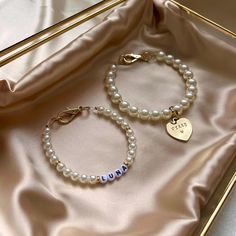 two bracelets with name charms on them sitting in a gold gift box next to a pink satin bag